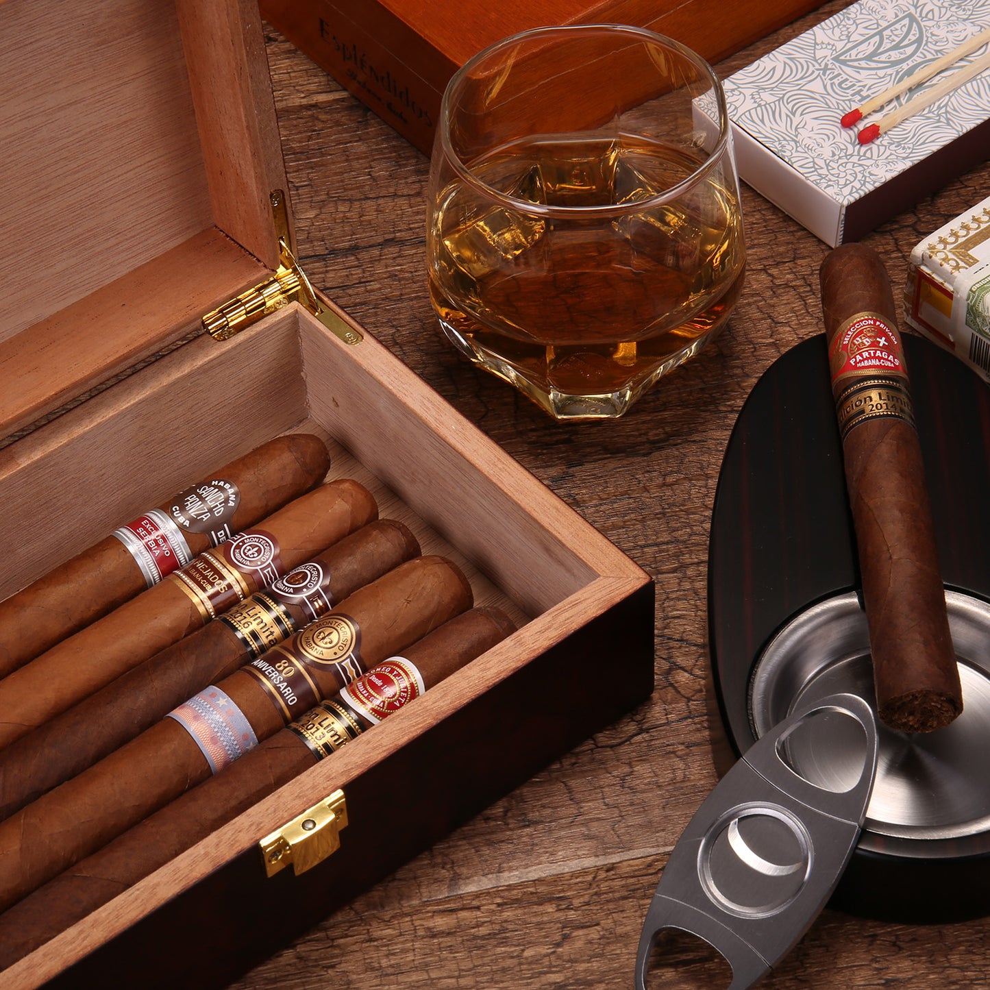 A close-up lifestyle shot of an open dark wooden cigar humidor filled with premium cigars, styled with a crystal glass of whiskey, a cigar resting on an ashtray, a cigar cutter, matchboxes, and a luxury cigar box in the background, all on a rustic wooden surface.