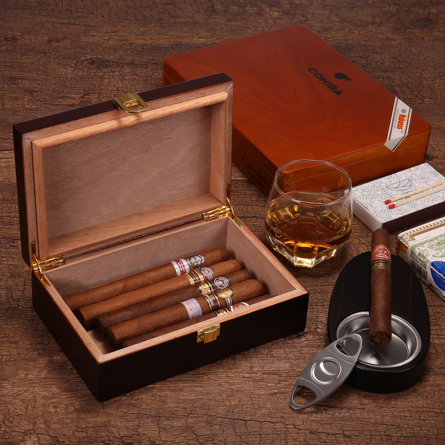 A lifestyle shot of an open cigar humidor filled with cigars, styled with a glass of whiskey, a cigar ashtray, a cigar cutter, matchboxes, and a closed COHIBA humidor in the background, all on a rustic wooden surface.
