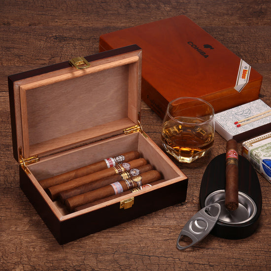 A lifestyle shot of an open cigar humidor filled with cigars, styled with a glass of whiskey, a cigar ashtray, a cigar cutter, matchboxes, and a closed COHIBA humidor in the background, all on a rustic wooden surface.