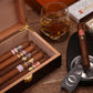 A close-up lifestyle shot of an open dark wooden cigar humidor filled with premium cigars, styled with a crystal glass of whiskey, a cigar resting on an ashtray, a cigar cutter, matchboxes, and a luxury cigar box in the background, all on a rustic wooden surface.