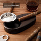A lifestyle shot of a solid wood cigar ashtray with a stainless steel bowl, holding a cigar, paired with a glass of whiskey and additional cigars, labeled "SOLID WOOD ASHTRAY" on a rustic wooden surface.