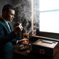 A moody luxury lifestyle shot of a man in a tailored suit smoking a cigar and holding a whiskey glass, with the humidor, ashtray, cutter, and accessories arranged on a wooden desk by a window, surrounded by cigar smoke.