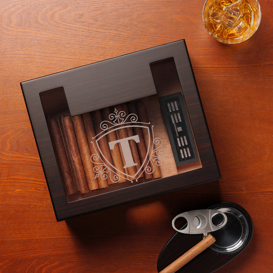 A custom dark wooden cigar humidor with a glass top, featuring a shield-style monogram "T" with decorative scrollwork, paired with a whiskey glass, cigar cutter, and ashtray on a textured wooden surface.