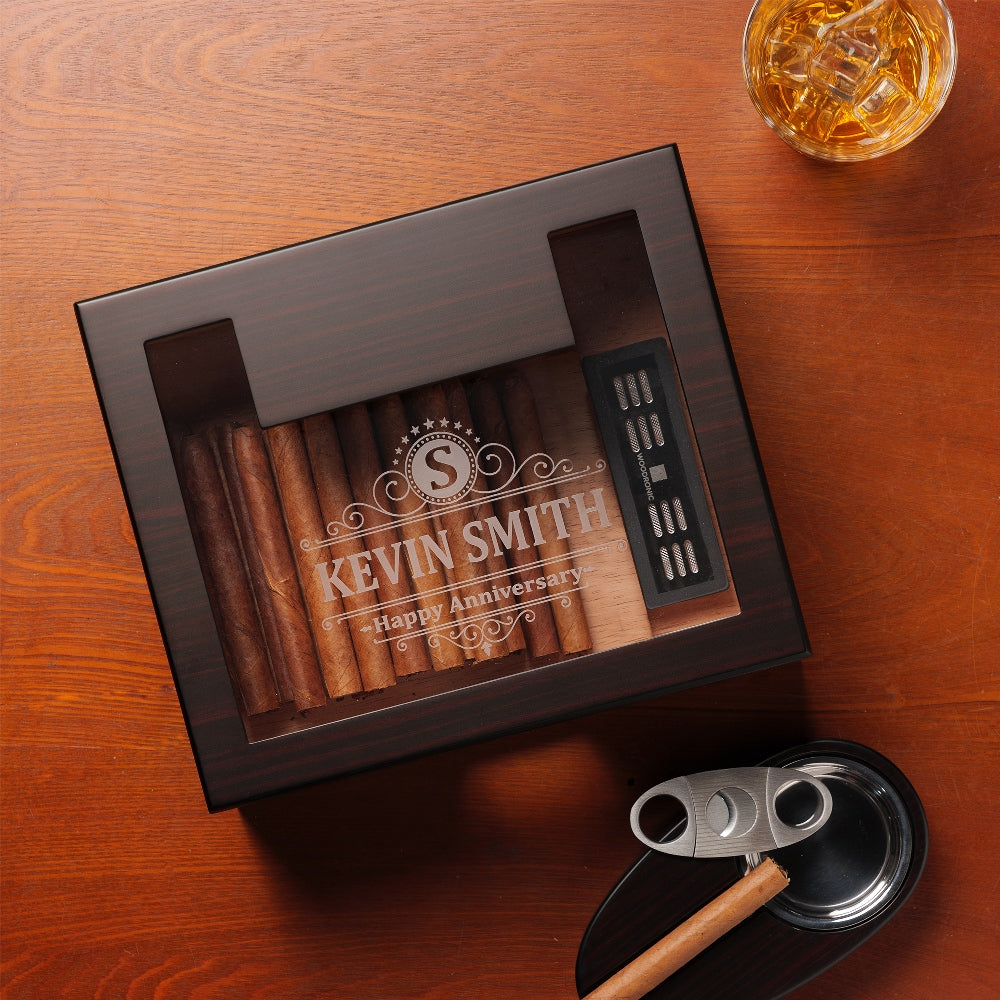 A personalized dark wooden cigar humidor with a glass top, engraved with "KEVIN SMITH - Happy Anniversary" and a monogram "S", styled with a glass of whiskey, cigar cutter, and ashtray on a rustic wooden table.