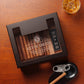 A personalized dark wooden cigar humidor with a glass top, engraved with "KEVIN SMITH - Happy Anniversary" and a monogram "S", styled with a glass of whiskey, cigar cutter, and ashtray on a rustic wooden table.