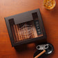 A custom dark wooden cigar humidor with a glass top, featuring "KEVIN SMITH" in a decorative banner design with ornate scrollwork, paired with a whiskey glass, cigar cutter, and ashtray on a textured wooden surface.