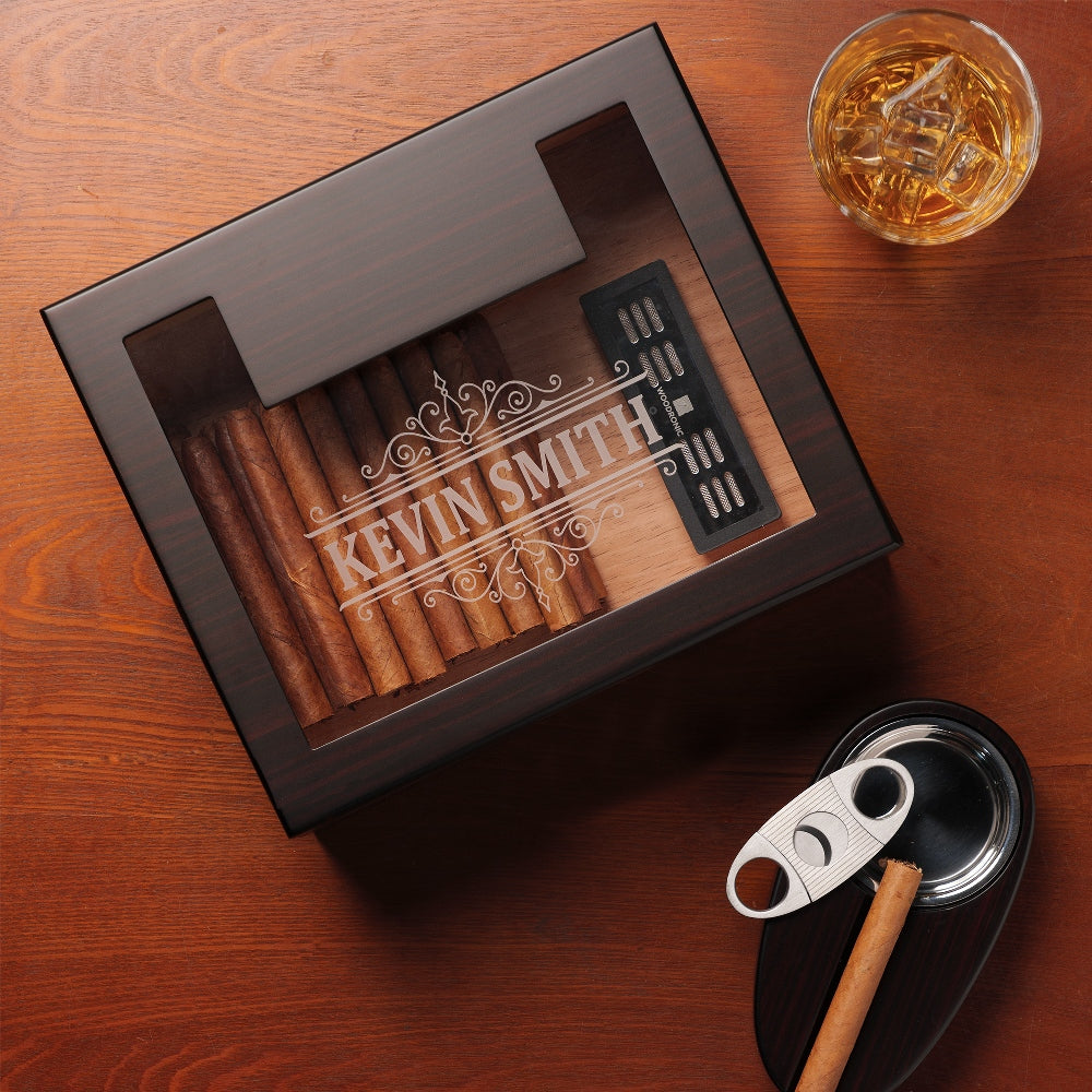 A personalized dark wooden cigar humidor with a glass top, engraved with "KEVIN SMITH" in an elaborate scroll font, accompanied by a glass of whiskey, cigar cutter, and ashtray on a rustic wooden table.