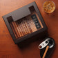 A personalized dark wooden cigar humidor with a glass top, engraved with "KEVIN SMITH" in an elaborate scroll font, accompanied by a glass of whiskey, cigar cutter, and ashtray on a rustic wooden table.