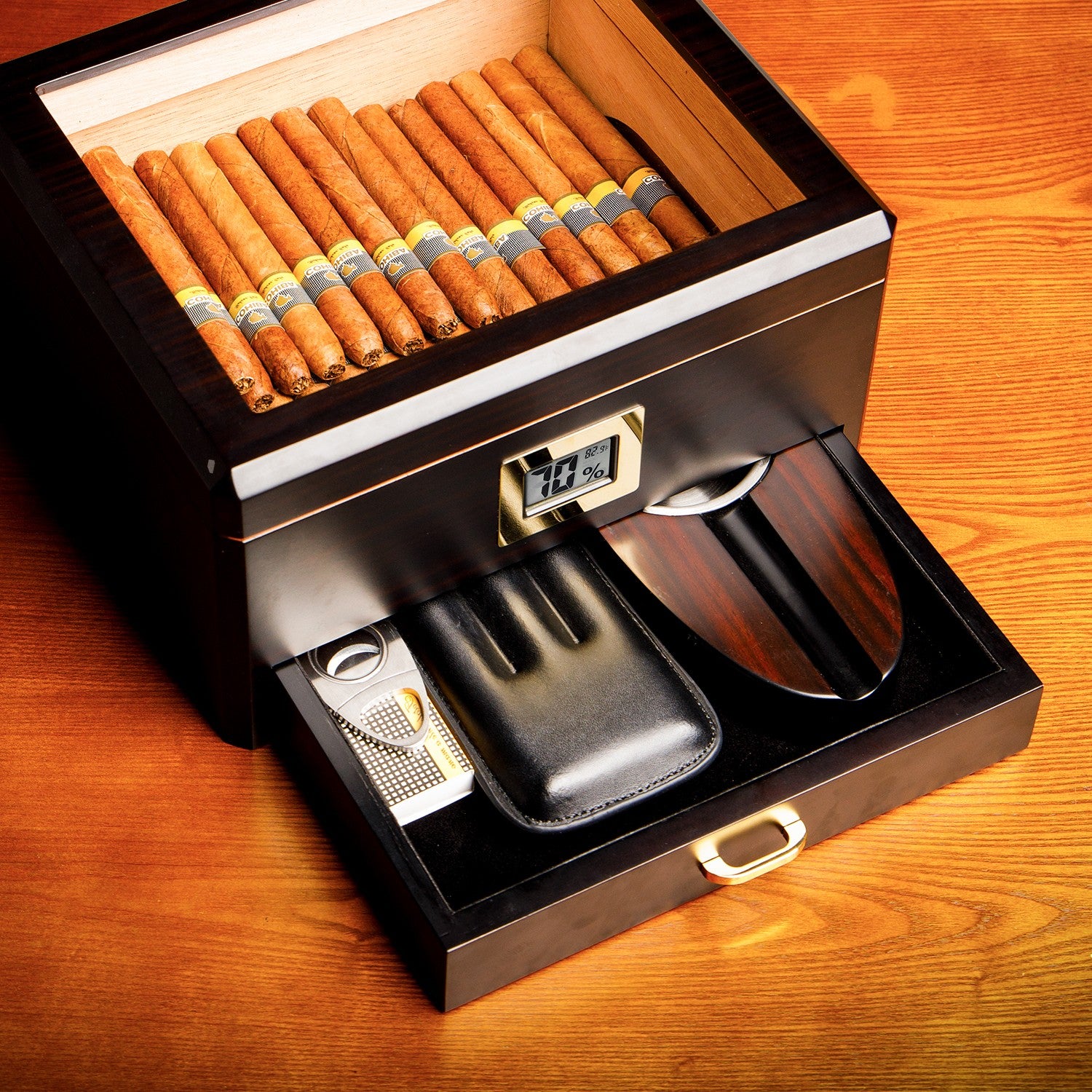 An open humidor filled with premium cigars, showing the pull-out accessory drawer containing a cigar cutter, leather case, and ashtray, placed on a warm wooden surface.