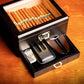 An open humidor filled with premium cigars, showing the pull-out accessory drawer containing a cigar cutter, leather case, and ashtray, placed on a warm wooden surface.
