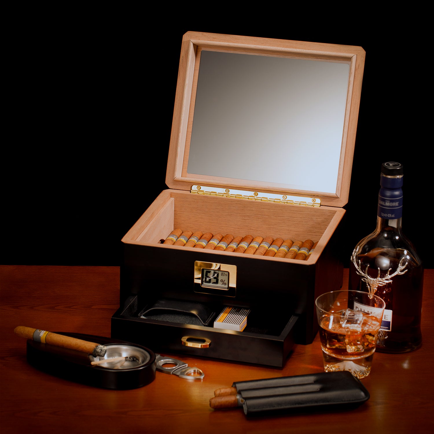 A sophisticated lifestyle scene featuring the open humidor filled with cigars, paired with a Dalmore whiskey bottle, glass, ashtray, cutter, and cigar case, on a wooden table.