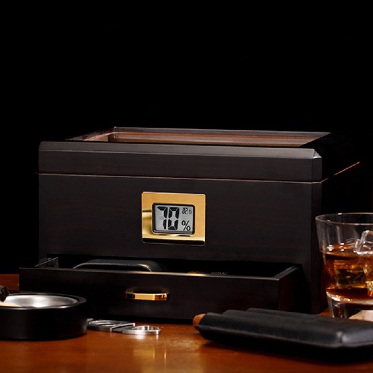 A moody lifestyle shot of the dark wood humidor with a digital hygrometer, styled with a glass of whiskey, ashtray, cigar cutter, and cigar case on a wooden table, against a black background.
