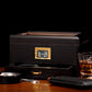 A moody lifestyle shot of the dark wood humidor with a digital hygrometer, styled with a glass of whiskey, ashtray, cigar cutter, and cigar case on a wooden table, against a black background.