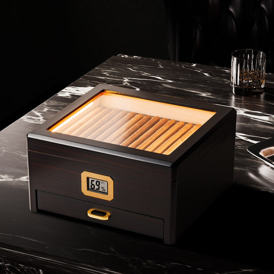 A sophisticated lifestyle shot of the LED-lit humidor placed on a black marble table, paired with a crystal whiskey glass and ashtray, creating an elegant premium aesthetic for cigar enthusiasts.