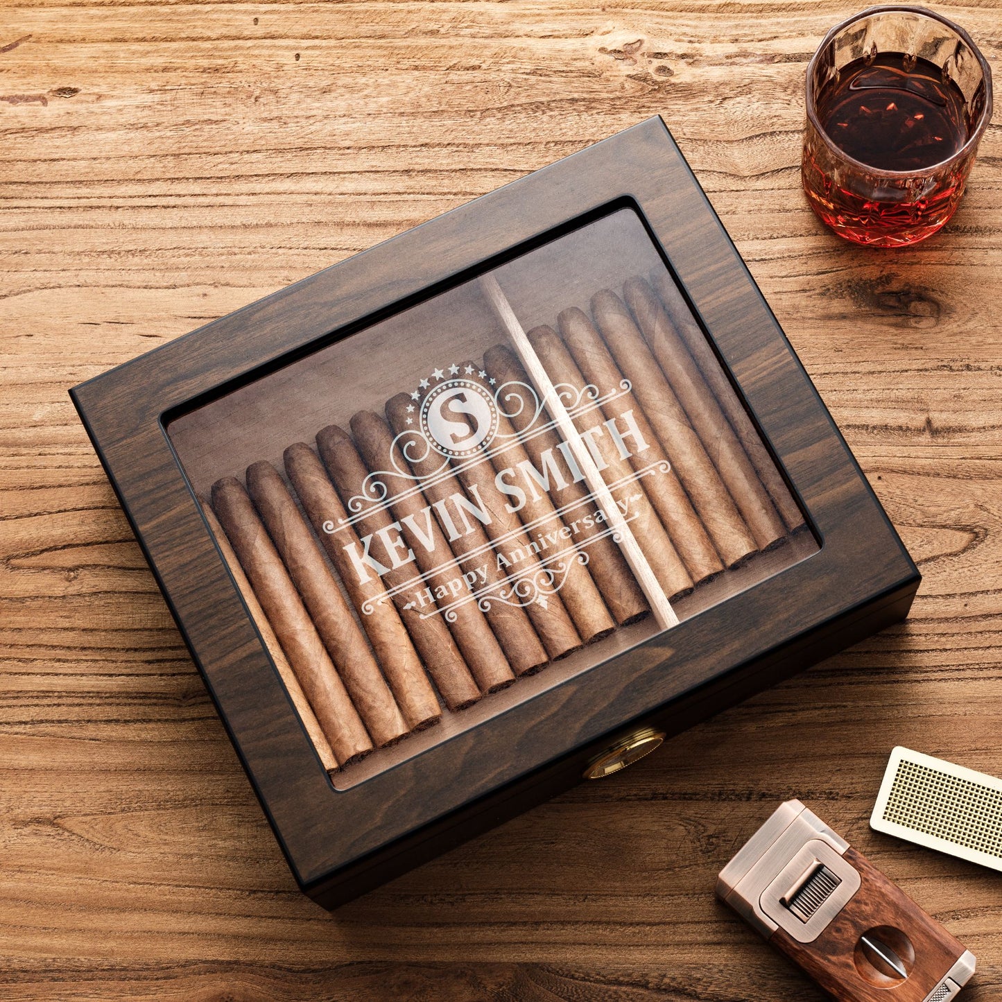 A personalized dark wooden cigar humidor with a glass top, engraved with "KEVIN SMITH - Happy Anniversary" and a monogram "S", styled with a glass of whiskey, cigar cutter, and humidifier bar on a rustic wooden table.