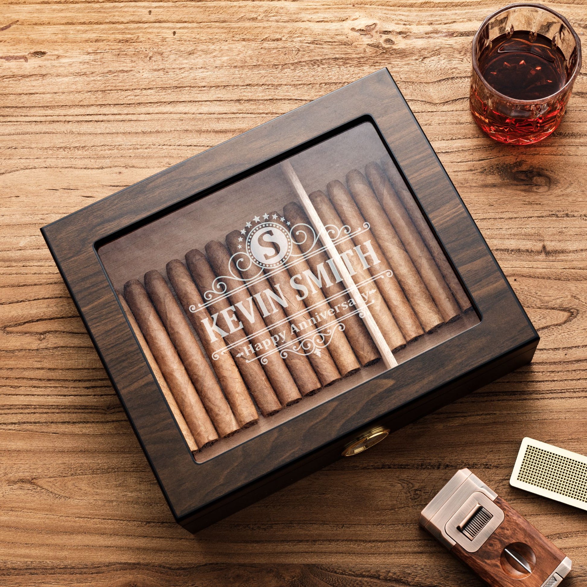 A personalized dark wooden cigar humidor with a glass top, engraved with "KEVIN SMITH - Happy Anniversary" and a monogram "S", styled with a glass of whiskey, cigar cutter, and humidifier bar on a rustic wooden table.