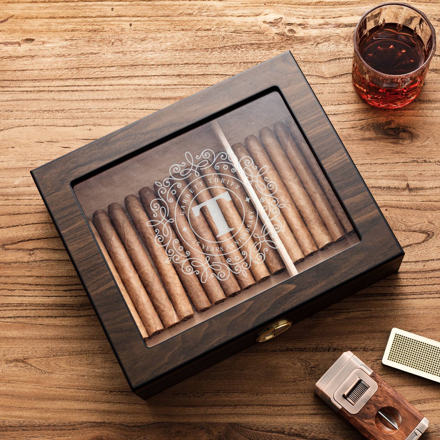 A custom dark wooden cigar humidor with a glass top, featuring a monogram "T" and anniversary text "GARRETT THRIFT 25 YEARS ANNIVERSARY", paired with a whiskey glass, cigar cutter, and humidifier bar on a textured wooden surface.