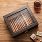 A personalized dark wooden cigar humidor with a glass top, engraved with "KEVIN SMITH" in an ornate scroll font, accompanied by a whiskey glass, cigar cutter, and humidifier bar on a textured wooden surface.