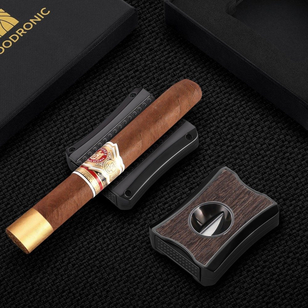 Two-tone wood veneer dual cigar cutter laid flat next to a full-size premium cigar, black fabric travel sleeve and branded gift box on textured black linen surface.