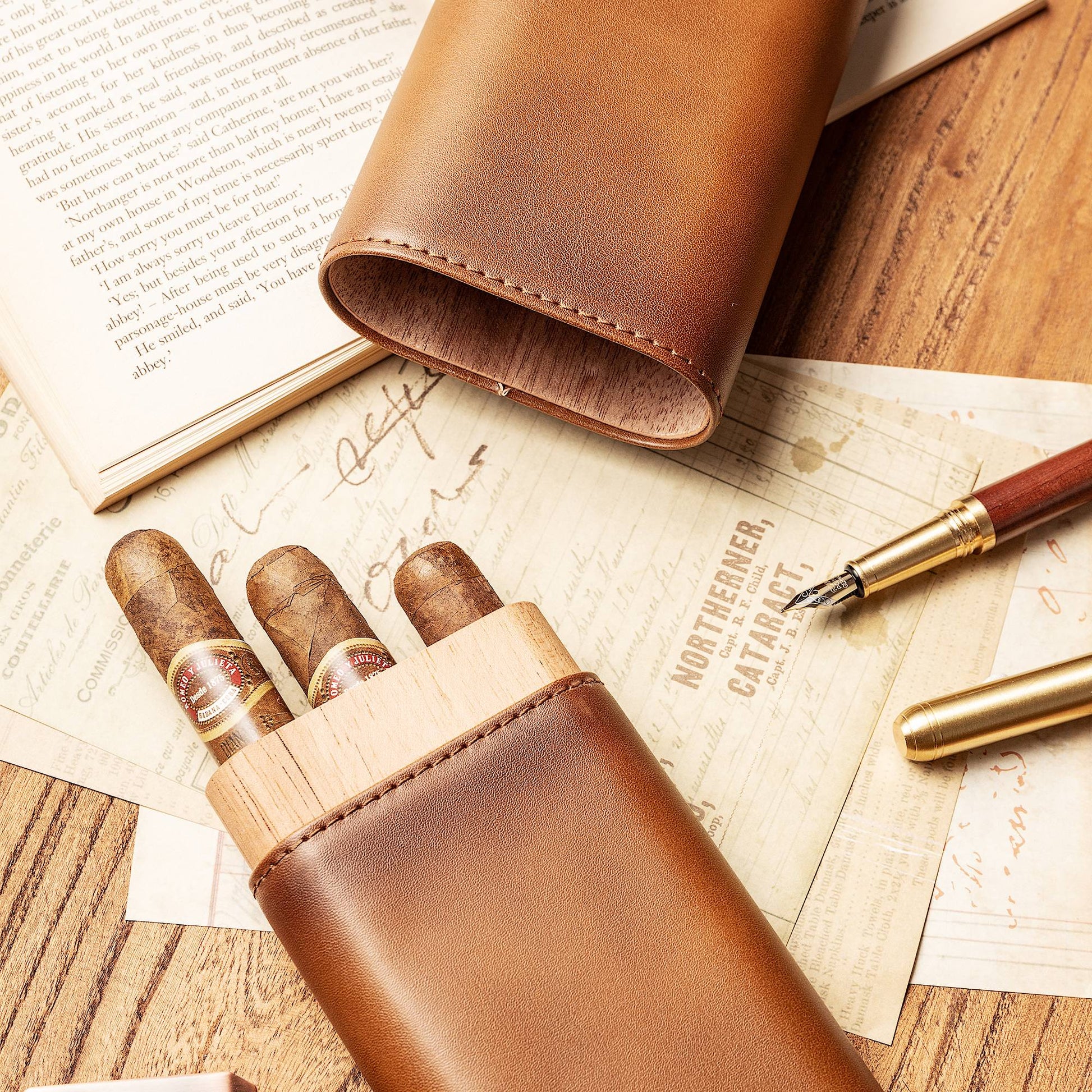 Smooth tan leather cigar case with exposed cedar wood top, holding three cigars, laid flat on aged documents alongside fountain pens, showcasing elegant craftsmanship.
