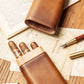 Smooth tan leather cigar case with exposed cedar wood top, holding three cigars, laid flat on aged documents alongside fountain pens, showcasing elegant craftsmanship.