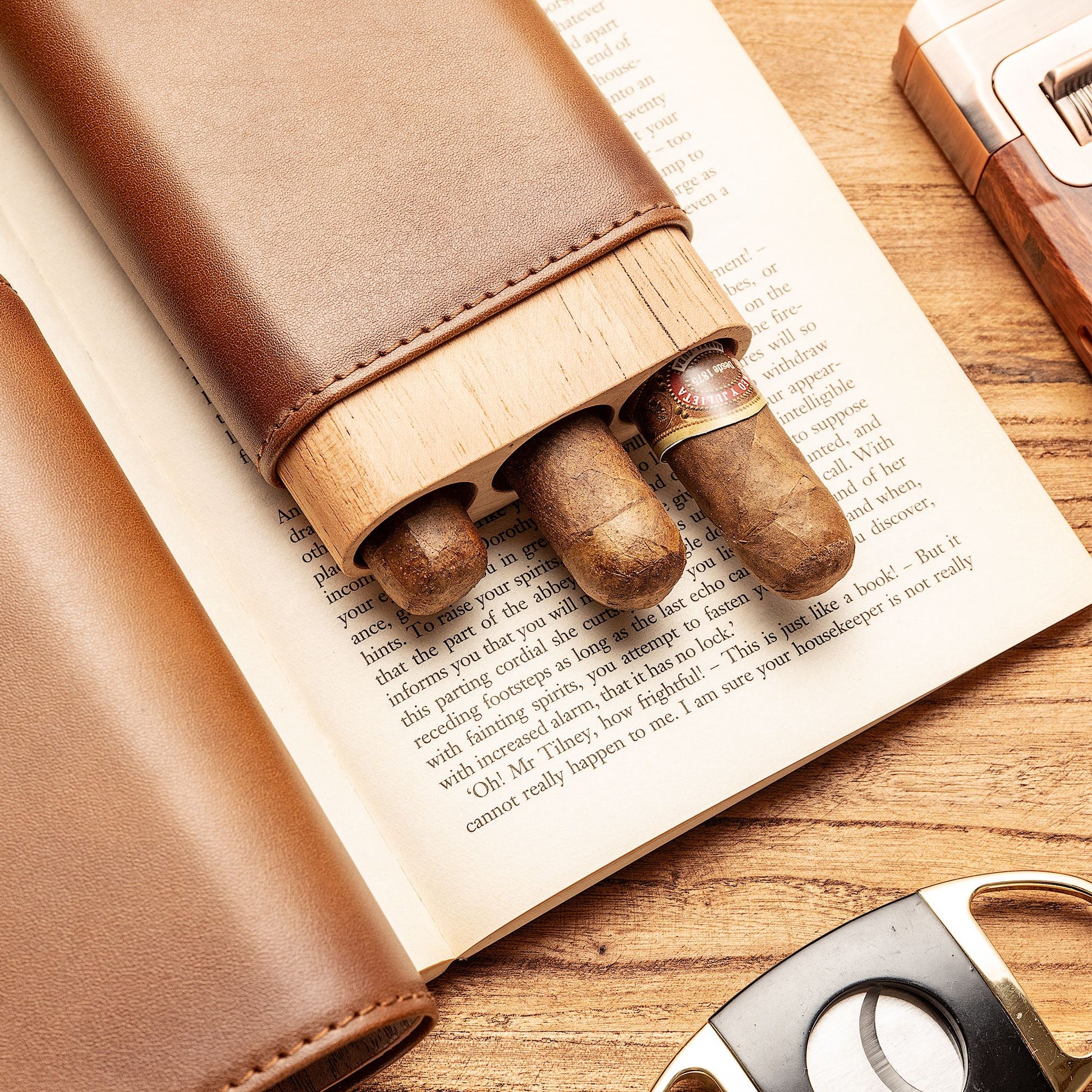 Leather cigar case with cedar liner, three cigars partially extended from its slots, resting on an open book with a cigar cutter nearby, highlighting on-the-go functionality.