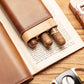 Leather cigar case with cedar liner, three cigars partially extended from its slots, resting on an open book with a cigar cutter nearby, highlighting on-the-go functionality.