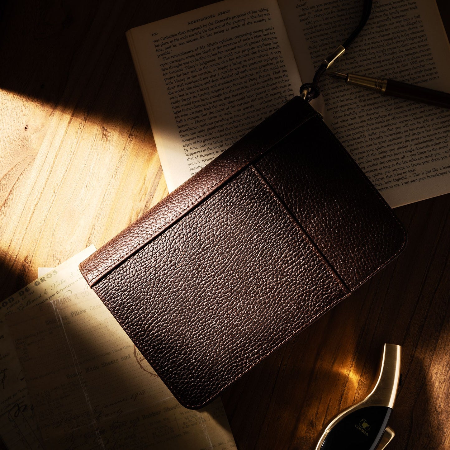 Closed dark brown leather cigar case resting on an open book in warm sunlight, with a fountain pen nearby, emphasizing its elegant, portable design for cigar enthusiasts.