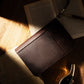 Closed dark brown leather cigar case resting on an open book in warm sunlight, with a fountain pen nearby, emphasizing its elegant, portable design for cigar enthusiasts.